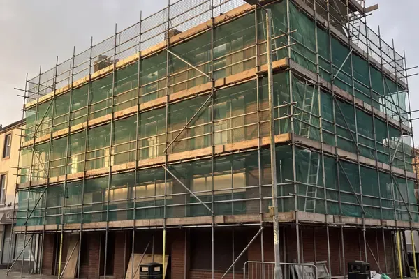 A three-storey corner commercial building sheeted in green debris netting, scaffolded to roof level above a street frontage.
