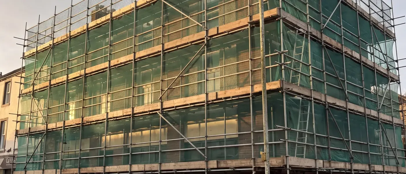 A three-storey corner commercial building sheeted in green debris netting, scaffolded to roof level above a street frontage.
