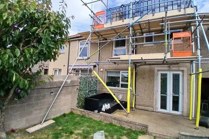 Scaffold to the side elevation of a rendered house, orange debris netting on the top lift and yellow-sleeved standards at ground level.