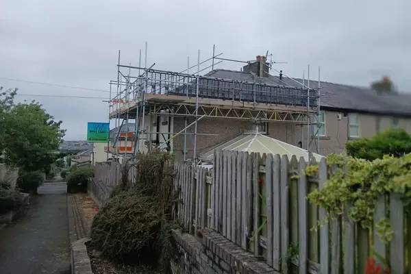 Boarded top lift running along a roof edge behind a garden fence, guardrails up and the fells behind.