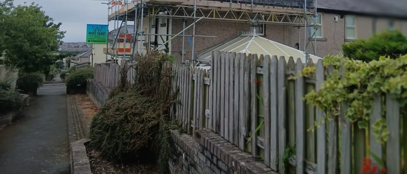 Boarded top lift running along a roof edge behind a garden fence, guardrails up and the fells behind.