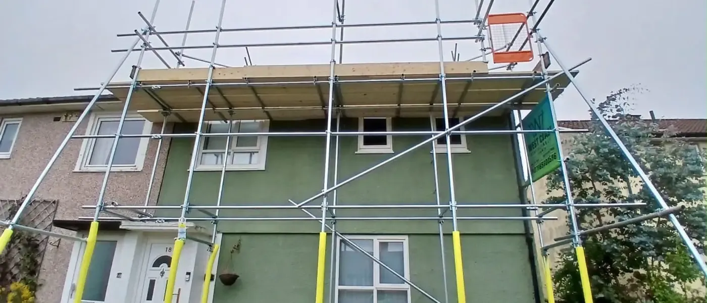 Scaffold to a green-rendered semi, boarded lift at eaves level with guardrails and yellow-sleeved standards.