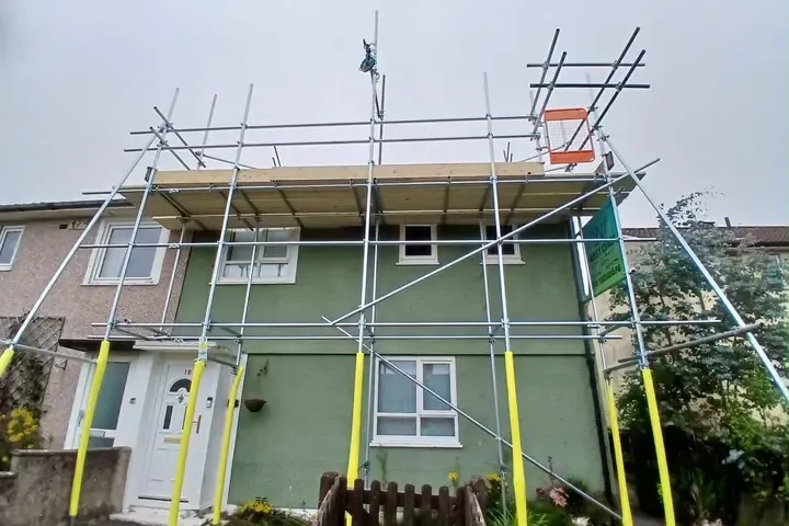 Scaffold to a green-rendered semi, boarded lift at eaves level with guardrails and yellow-sleeved standards.