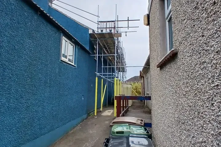 Scaffold standards running up a narrow passage between two houses, boarded lift above.