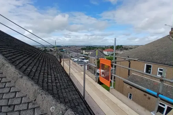 Scaffold at roof level with guardrails and an orange brick-guard panel, looking out over the rooftops toward the coast.