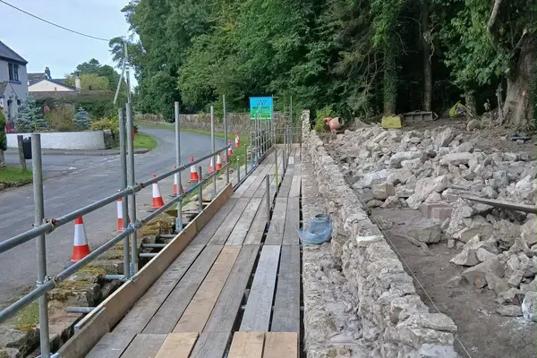 Boarded pedestrian walkway with cones and a handrail, carrying the footway past a rebuilt stone wall.