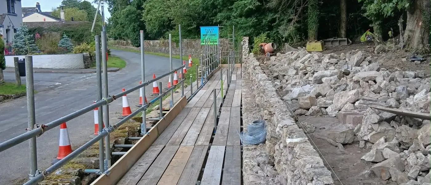 Boarded pedestrian walkway with cones and a handrail, carrying the footway past a rebuilt stone wall.