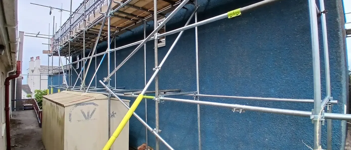 Scaffold standards against a blue-painted gable, boarded lift running above a yard.