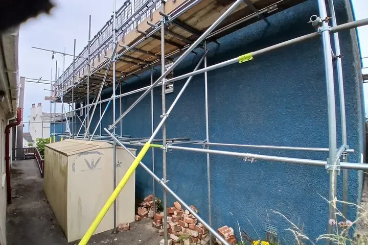 Scaffold standards against a blue-painted gable, boarded lift running above a yard.
