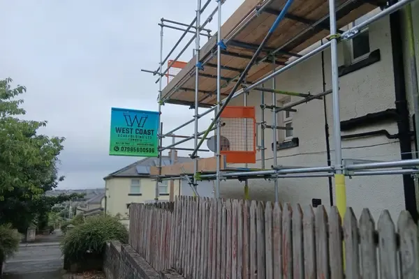 Scaffold to a gable end with the company banner tied on, boarded lift and orange netting above a fenced yard.