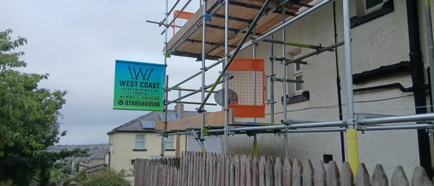 Scaffold to a gable end with the company banner tied on, boarded lift and orange netting above a fenced yard.