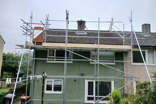 Scaffold to a green-rendered house, two boarded lifts and yellow-sleeved standards along the drive.