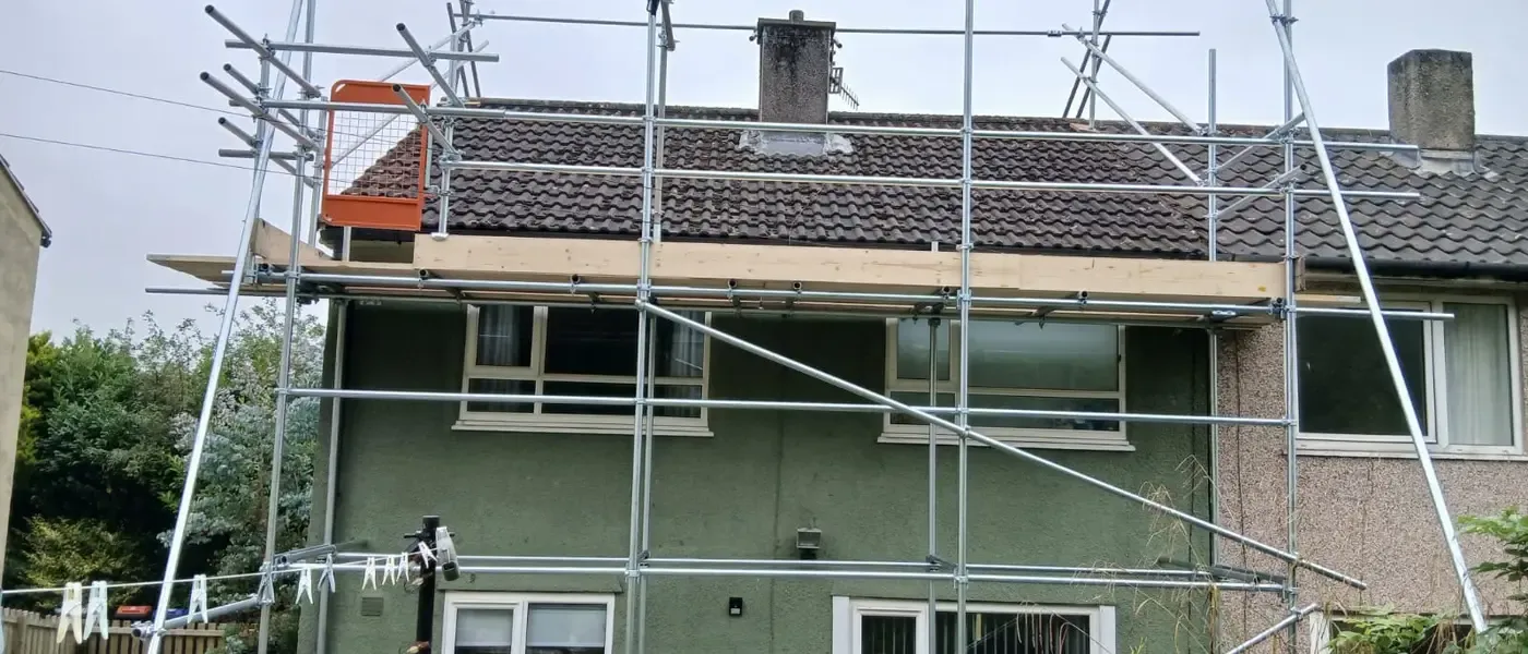 Scaffold to a green-rendered house, two boarded lifts and yellow-sleeved standards along the drive.